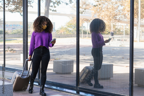 Young woman in protective face mask using smart phone while walking with luggage by glass photo