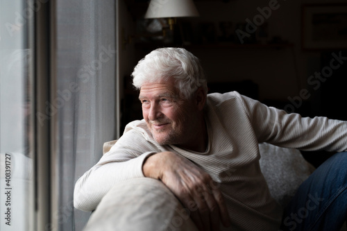 Smiling senior man looking through window while sitting on sofa at home