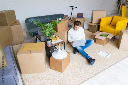 Young many using laptop in messy living room while moving in new home photo
