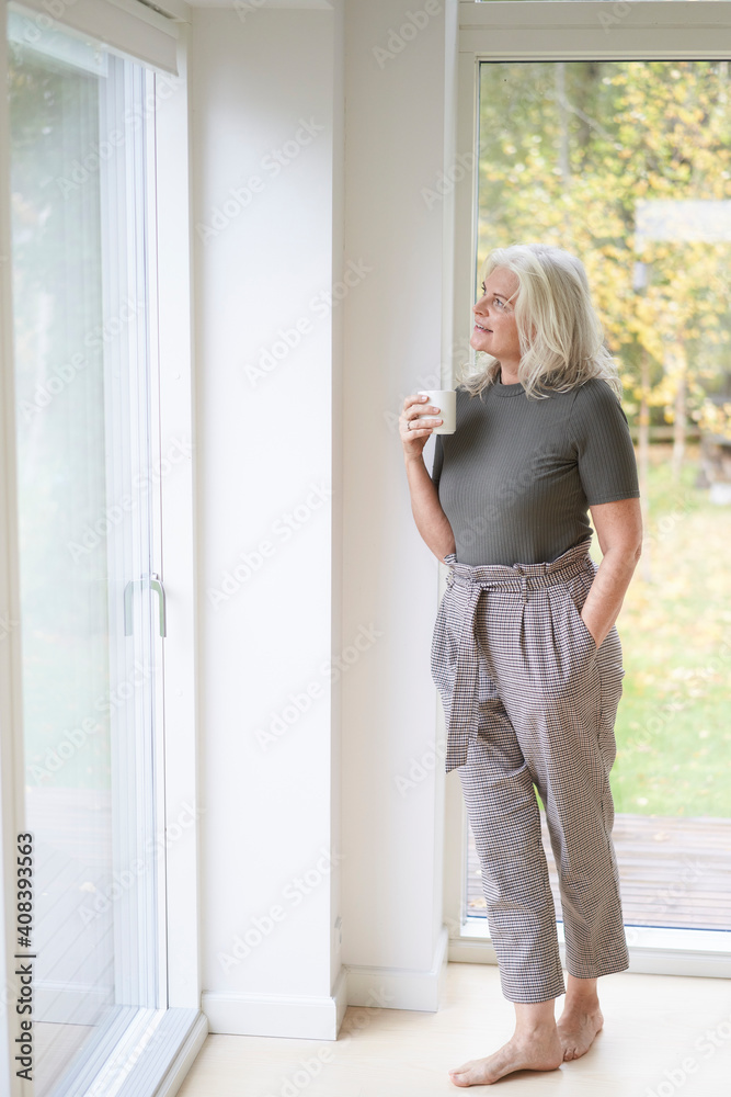 Thoughtful retired woman holding cup while looking through window at home Thoughtful retired woman holding cup while looking through window at home