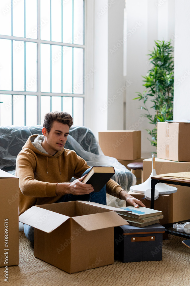 Young man unpacking books from box while moving in new loft apartment Young man unpacking books from box while moving in new loft apartment