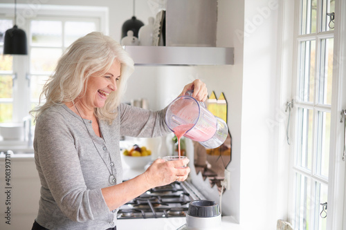 Cheerful senior woman pouring strawberry smoothie in glass at kitchen