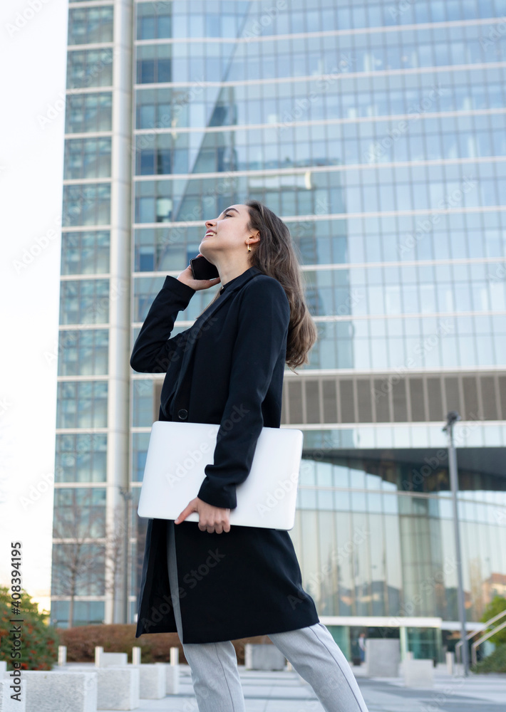 Businesswoman with laptop smiling while talking on mobile phone walking in city Businesswoman with laptop smiling while talking on mobile phone walking in city