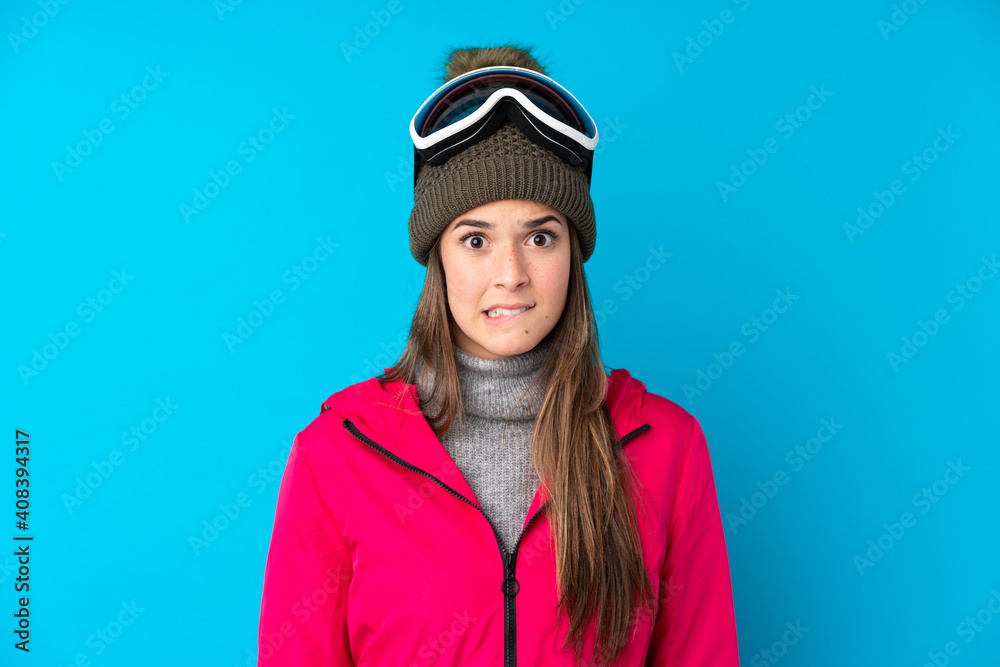 Teenager skier girl with snowboarding glasses over isolated blue background having doubts and with confuse face expression