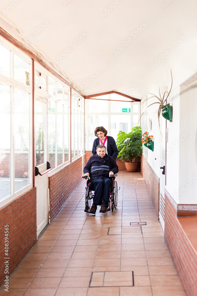 Female nurse helping disabled man sitting on wheelchair in corridor of nursing home Female nurse helping disabled man sitting on wheelchair in corridor of nursing home