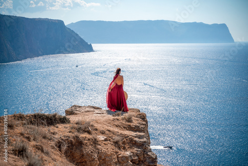 A girl with loose hair in a long red dress descends the stairs between the yellow rocks overlooking the sea. A rock can be seen in the sea. Sunny path on the sea from the rising sun © svetograph