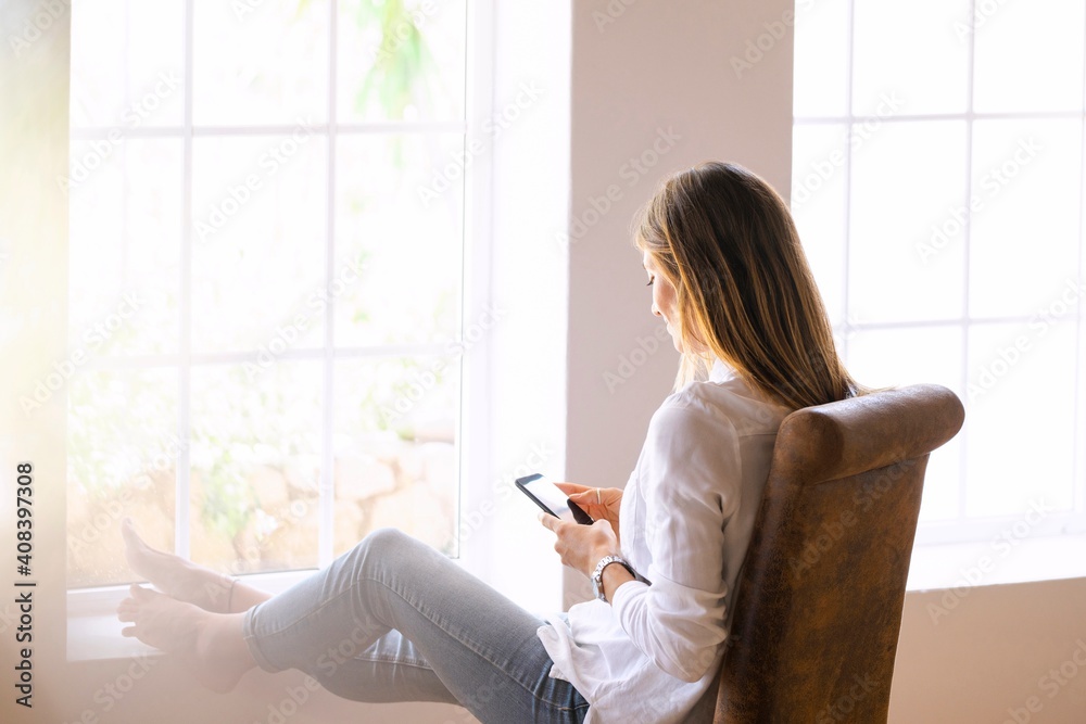 Young woman using smart phone while sitting on chair relaxing by window at home Young woman using smart phone while sitting on chair relaxing by window at home