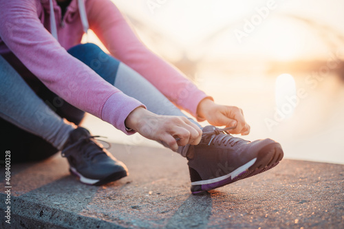 Close up shot of female jogger ties shoes on sneakers. Beautiful river bridge and sunset in the background.