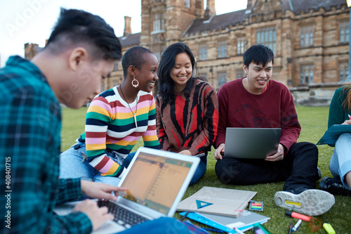Group of young university students hanging out sitting on grass studying and using devices photo