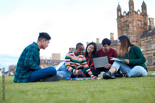 Group of young university students hanging out sitting on grass studying and using devices photo