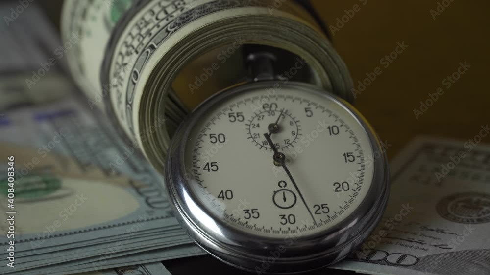 The stopwatch is on the table, next to it is a wad of 100 dollars bills rolled up with a rubber band, and the hand is ticking. The concept of sales, discounts and investments in stocks and businesses. The stopwatch is on the table, next to it is a wad of 100 dollars bills rolled up with a rubber band, and the hand is ticking. The concept of sales, discounts and investments in stocks and businesses.