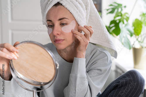 Tableau sur toile Woman with towel on head applying patches under eyes enriched with collagen, vitamin E, diminishes the signs of aging, helps reduse eye puffiness, looking in mirror