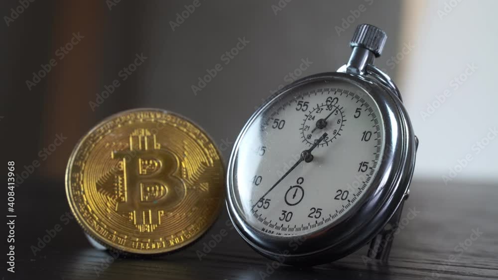 A gold bitcoin and an analog metal stopwatch are lying side by side on a black table. Close-up. A gold bitcoin and an analog metal stopwatch are lying side by side on a black table. Close-up.