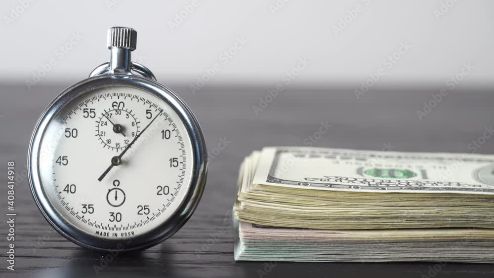 The stopwatch is on the table, next to it is a wad of 100 dollars bills, and the hand is ticking. The concept of sales, discounts and investments in stocks and businesses. The stopwatch is on the table, next to it is a wad of 100 dollars bills, and the hand is ticking. The concept of sales, discounts and investments in stocks and businesses.