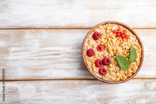 Wheat flakes porridge with milk, raspberry and currant in wooden bowl on white wooden background. Top view, copy space.