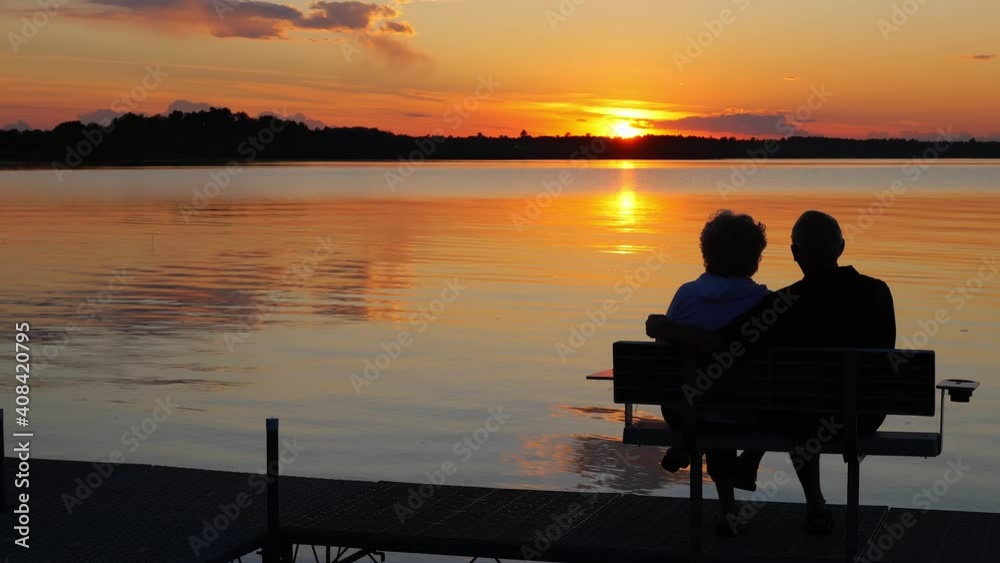 Silhouette of two people on a bench on a dock as they enjoy a sunset over a beautiful lake in Minnesota on a serene and relaxing evening, in golden hour light.