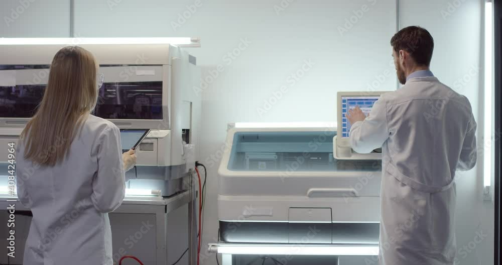 Back view of Caucasian male and female doctors professionals standing in laboratory working and speaking. Man specialist using medical equipment computer for diagnostic, woman typing on tablet Back view of Caucasian male and female doctors professionals standing in laboratory working and speaking. Man specialist using medical equipment computer for diagnostic, woman typing on tablet