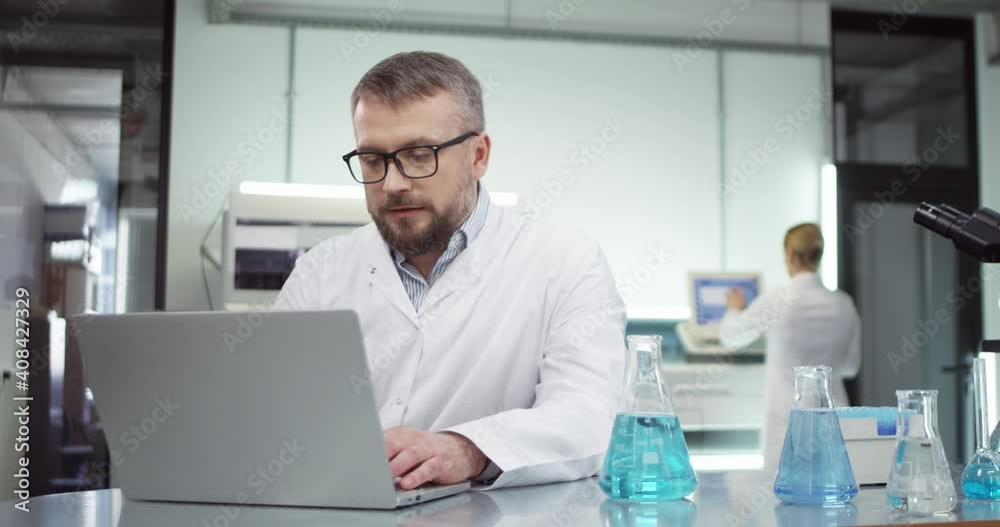 Portrait of busy Caucasian middle-aged professional male doctor scientist browsing online on laptop computer working in hospital laboratory with many tubes with liquid on table, vaccine development