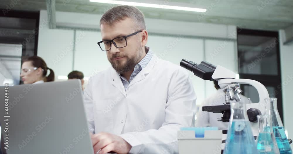 Portrait of happy middle-aged Caucasian male doctor laboratory scientist sitting in lab and texting browsing online on laptop, checking analysis and vaccine results, medical research, hospital concept Portrait of happy middle-aged Caucasian male doctor laboratory scientist sitting in lab and texting browsing online on laptop, checking analysis and vaccine results, medical research, hospital concept