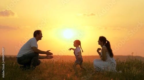 Happy family walks in park at sunset. Mom, dad and baby. Little daughter goes from mom to dad, hugs and kisses her parents in rays of warm sun. Healthy family plays in field. Happy family concept