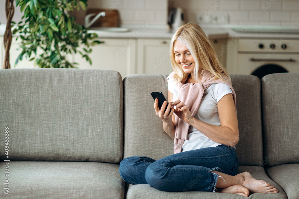 Mature blonde caucasian woman sits at the sofa, using smartphone. Happy middle aged woman chat with friends or family, browsing internet, smiling Mature blonde caucasian woman sits at the sofa, using smartphone. Happy middle aged woman chat with friends or family, browsing internet, smiling