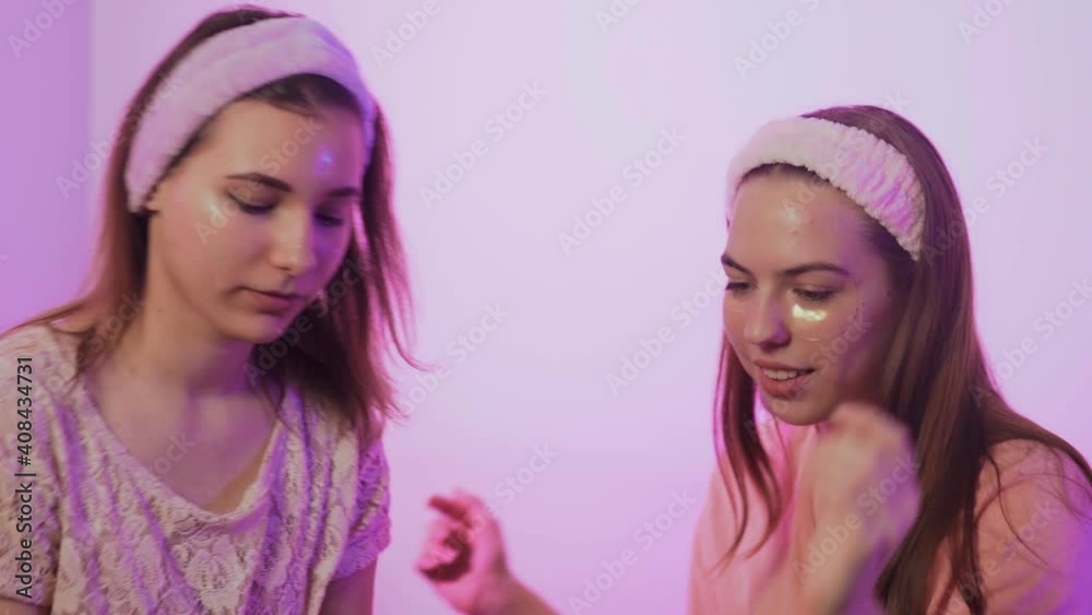 Two happy pretty young women in pajamas, Eye Collagen Patches and headbands on head sits on bed and takes photo on phone at bachelorette party Two happy pretty young women in pajamas, Eye Collagen Patches and headbands on head sits on bed and takes photo on phone at bachelorette party