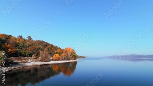 the footage of a seascape surrounded by mountains and colorful trees under the blue sky photo