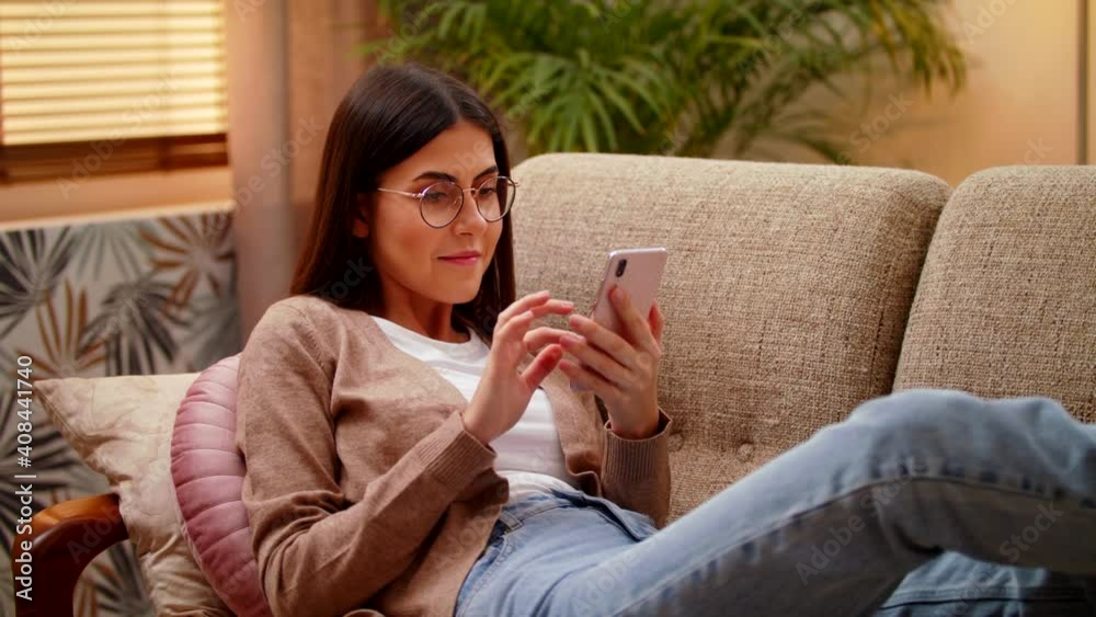 Attractive Smiling Young Woman Lying, Resting On A Sofa In The Bright Cozy Living Room At Home And Holding Smart Phone Watching And Chatting In Social Media Using Mobile Apps, Doing Shopping Attractive Smiling Young Woman Lying, Resting On A Sofa In The Bright Cozy Living Room At Home And Holding Smart Phone Watching And Chatting In Social Media Using Mobile Apps, Doing Shopping
