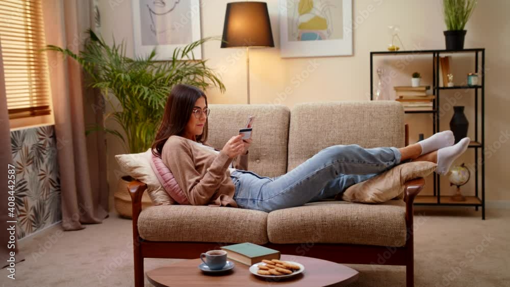 Smiling Woman Making Online Payment Holding Credit Card And Shopping Online Using Smartphone Lying On Sofa In Living Room At Home. Concept Of E-commerce, E-Banking, Finance, Shopping In Internet Smiling Woman Making Online Payment Holding Credit Card And Shopping Online Using Smartphone Lying On Sofa In Living Room At Home. Concept Of E-commerce, E-Banking, Finance, Shopping In Internet