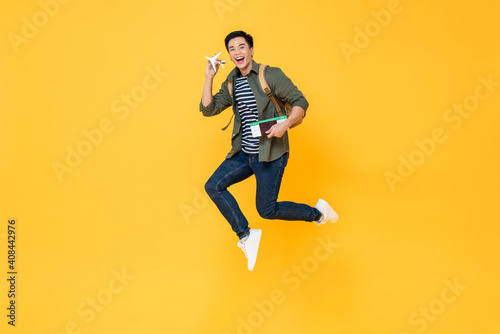 Young handsome Asian tourist man holding airplane model, passport and boarding pass jumping in isolated yellow color background