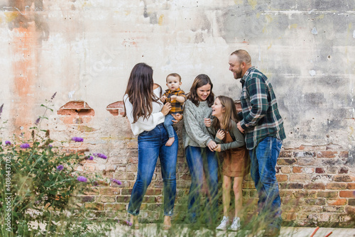 A family of five with two girls and a baby boy standing by an urban old brick wall and laughing and playing together