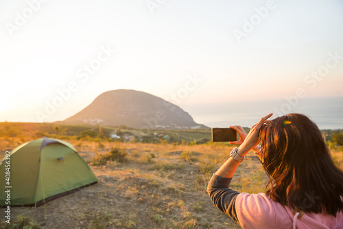 Woman takes photos of the sunrise in the mountains on her phone. Selfie in rising sun. Panoramic view on sea and Ayu-Dag. Camping  outdoor activities  sports mountain hiking  family travel. Crimea.