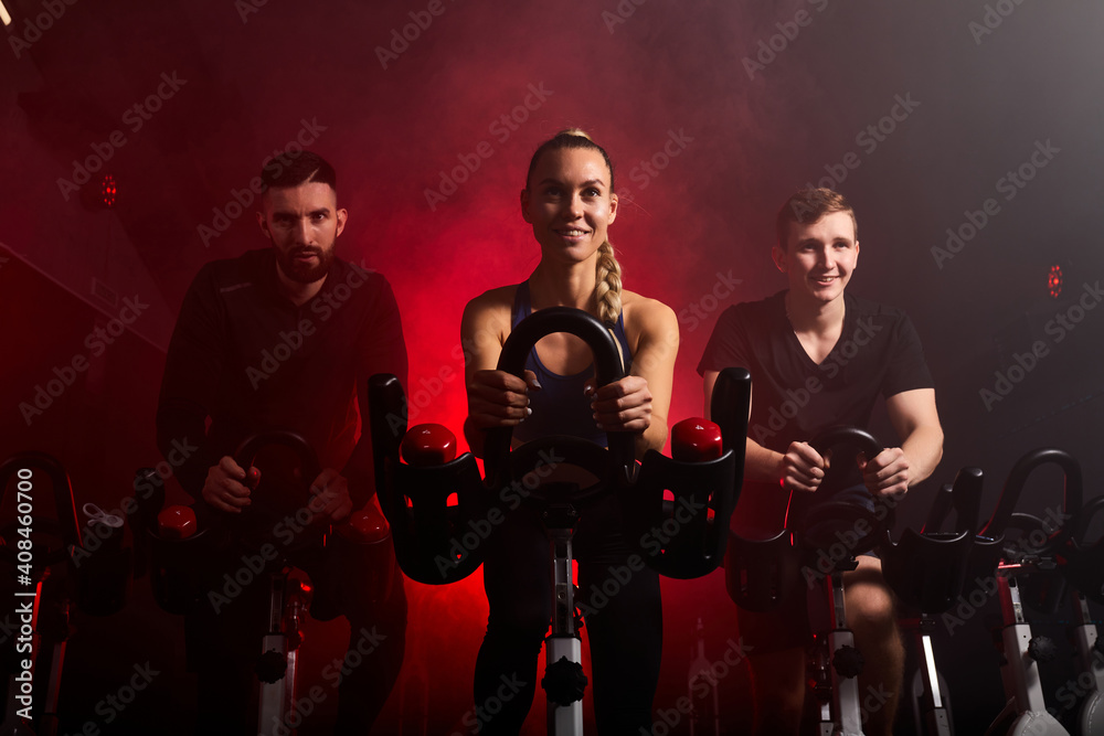 Fototapeta premium fit people riding exercise bikes in gym, enjoying workout time, in smoky red neon space