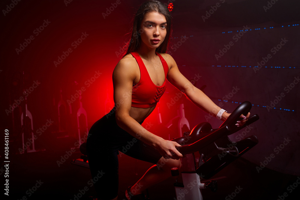 female in sportswear exercising on bicycle at modern gym, workout woman training on smart stationary bike indoors in red neon lighted smoky space