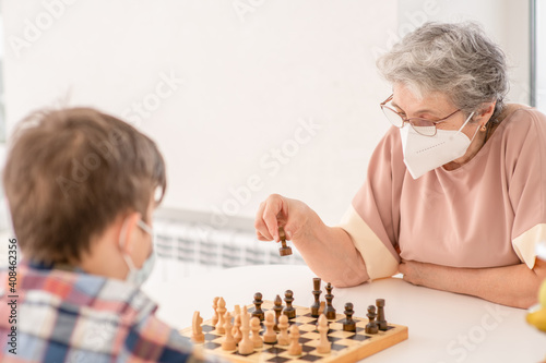 Senior woman and her grandson wearing protective face masks play chess at home during quarantine Coronavirus (Covid-19) epidemic