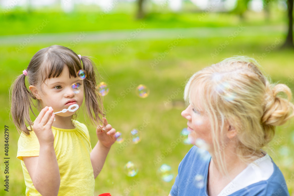 Little girl with syndrome down blows bubbles in a summer park with her mother