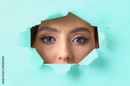 Face of beautiful young woman with blue eyes visible through hole in color paper © Pixel-Shot