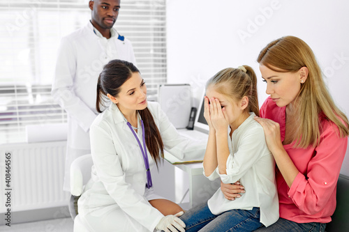 female doctor general practitioner talks and amuses child before medical examination, calms down the crying girl closing face with hands, came with mother