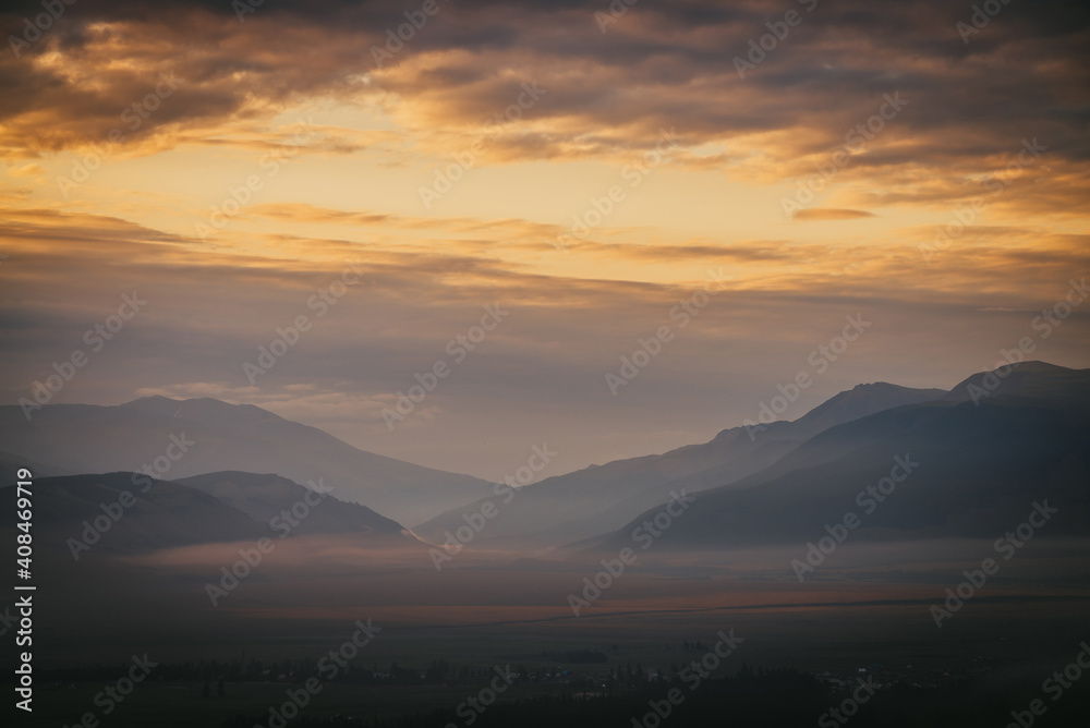 Scenic dawn mountain landscape with golden low clouds in valley among mountains silhouettes under cloudy sky. Vivid sunset or sunrise scenery with low clouds in mountain valley in illuminating color. Scenic dawn mountain landscape with golden low clouds in valley among mountains silhouettes under cloudy sky. Vivid sunset or sunrise scenery with low clouds in mountain valley in illuminating color.