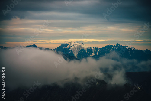Atmospheric dawn mountain landscape with great snowy mountain peak in sunlight above big low cloud among rocks and sunset skylight of illuminating color in cloudy sky. Illuminating sunlight in sunrise