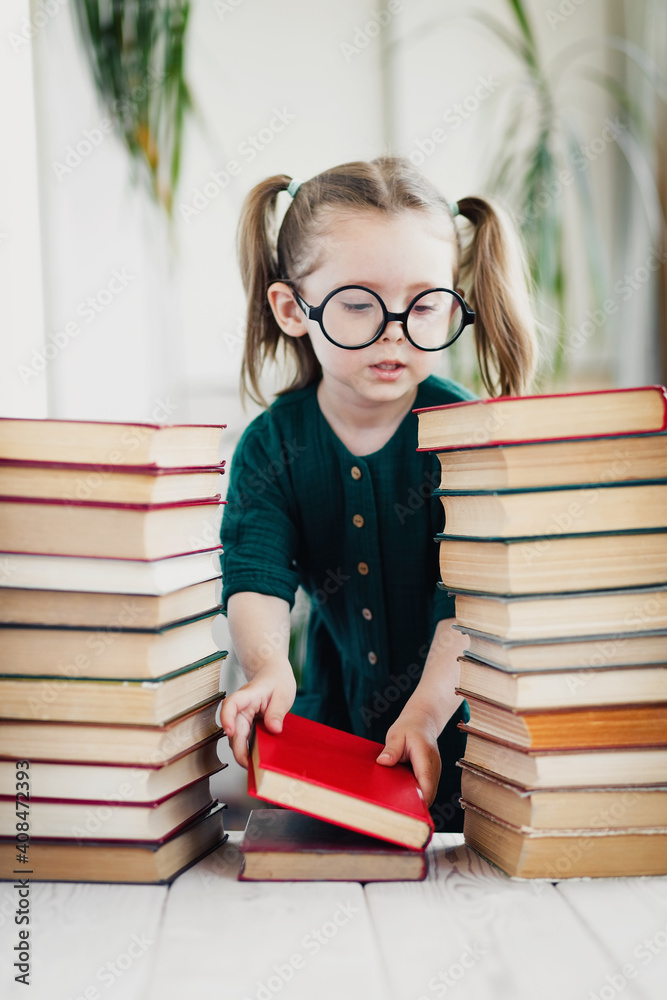 Cute little girl in round shaped glasses and green muslin dress folds colorful books in order. Cute little girl in round shaped glasses and green muslin dress folds colorful books in order.