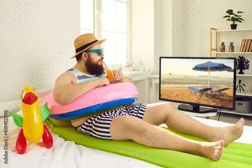 Funny young man with sunglasses and inflatable beach toys sipping cocktail and watching travel show on TV. Concept of canceled summer holiday plans, vacation in lockdown at home or Covid-19 quarantine