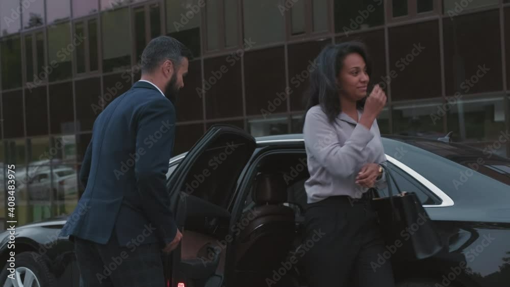 Medium shot of successful mixed race business lady getting out of backseat of expensive black car and male driver in suit opening car door for her