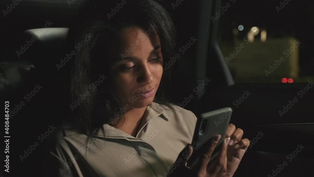 Close-up of adult mixed race business-like woman looking at smartphone screen smiling while sitting at backseat of posh car at night Close-up of adult mixed race business-like woman looking at smartphone screen smiling while sitting at backseat of posh car at night