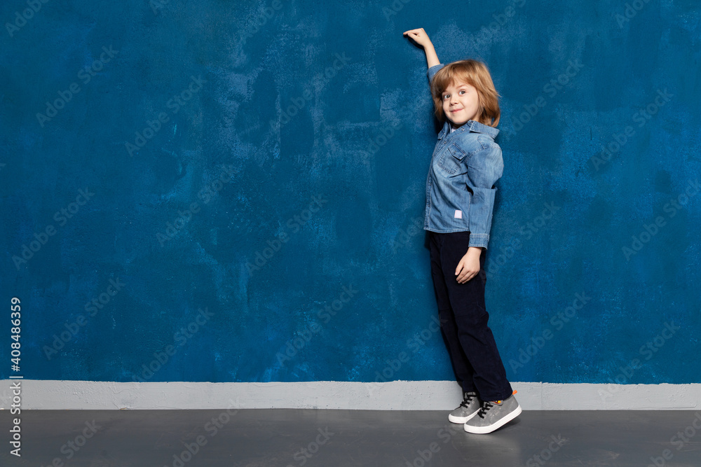 Smiling kid boy show height, measure tall growth by arm raised up on empty wall background with copyspace for advertising. Caucasian schoolboy in casual clothes demonstrates size on blank backdrop.