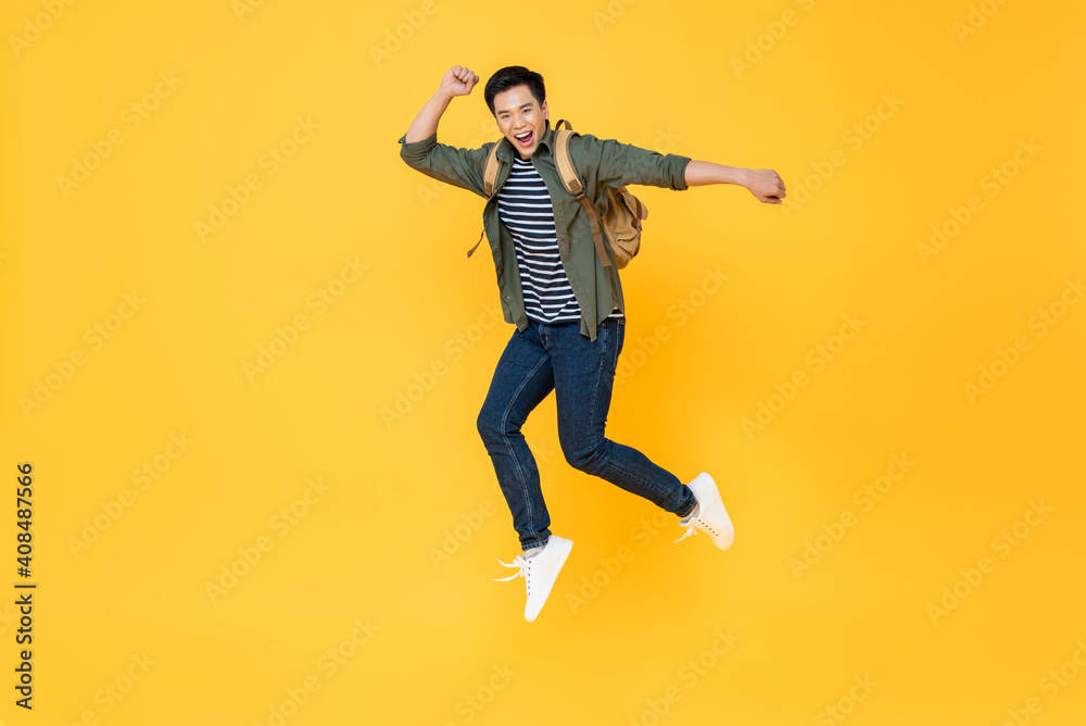 Happy smiling young Asian tourist man with backpack jumping in mid-air isolated on yellow background
