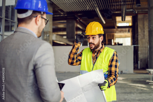 Construction worker holding blueprints and talking to his supervisor while standing at construction site.