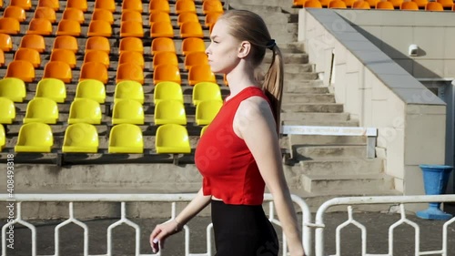 young slim busty jogger female in sportswear walking. fit caucasian pretty woman in red activewear walks outdoors along statium tribune before fitness training, workout. sports, healthy lifestyle photo