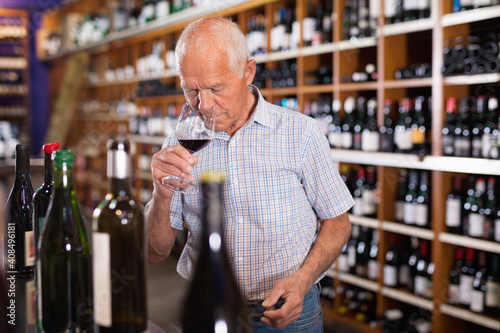 Confident elderly positive smiling man tasting red wine in wine store before buying.