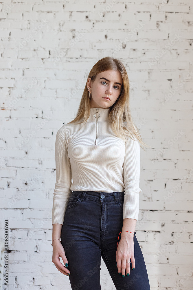 portrait of cute thoughtful teen model wearing white sweater and black jeans. caucasian skinny young female with blond hair stands in front of white brick wall. natural pretty lady standing at studio portrait of cute thoughtful teen model wearing white sweater and black jeans. caucasian skinny young female with blond hair stands in front of white brick wall. natural pretty lady standing at studio